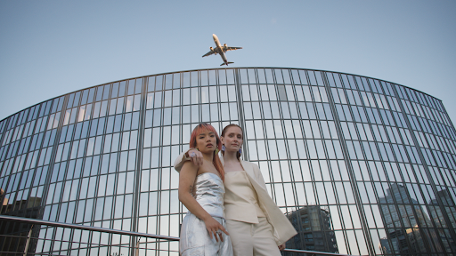 Zwei Frauen, die sich umarmen, stehen nebeneinander vor einem großen Gebäude mit vielen Fenstern. Im Hintergrund fliegt ein Flugzeug über das Gebäude.