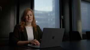 Szene Infofilm: Eine Frau schaut in die Kamera während sie interviewt wird. Vor ihr auf dem Tisch steht ein Laptop.