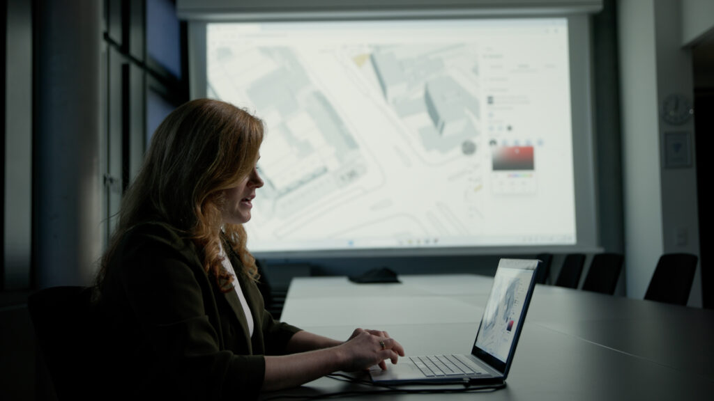 Eine Frau sitzt an einem Laptop in einem Konferenzraum. Hinter ihr ist unscharf eine große Projektion eines digitalen Stadtplans zu sehen.