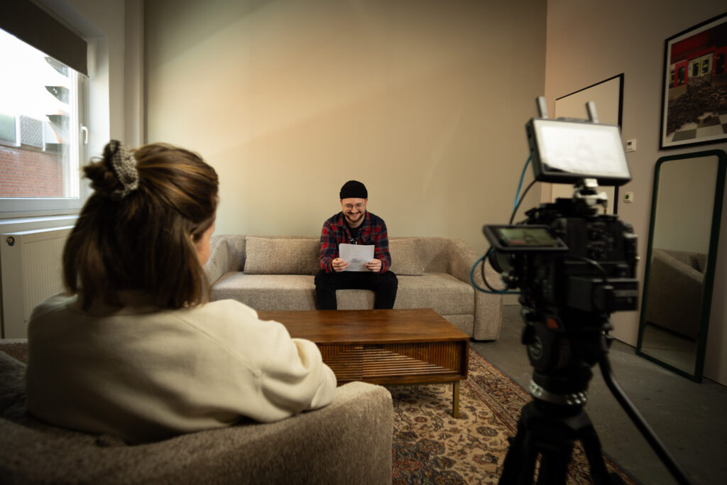 Ein Mann sitzt auf einem Sofa und hält einen weißen Zettel in der Hand. Er schaut zu einer Frau, die der Kamera den Rücken zu wendet. Das Gespräch der beiden wird mit einer Kamera aufgezeichnet.