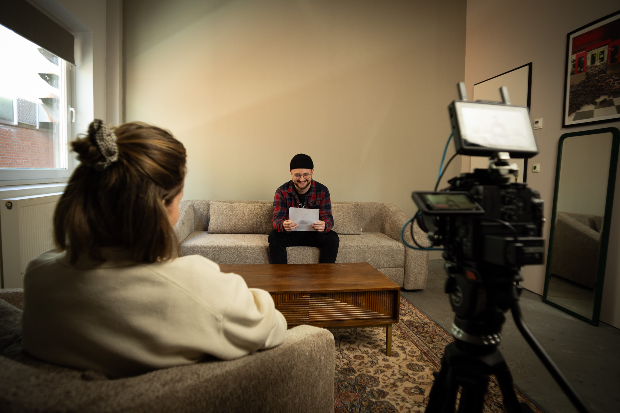 Ein Mann sitzt auf einem Sofa und hält einen weißen Zettel in der Hand. Er schaut zu einer Frau, die der Kamera den Rücken zu wendet. Das Gespräch der beiden wird mit einer Kamera aufgezeichnet.